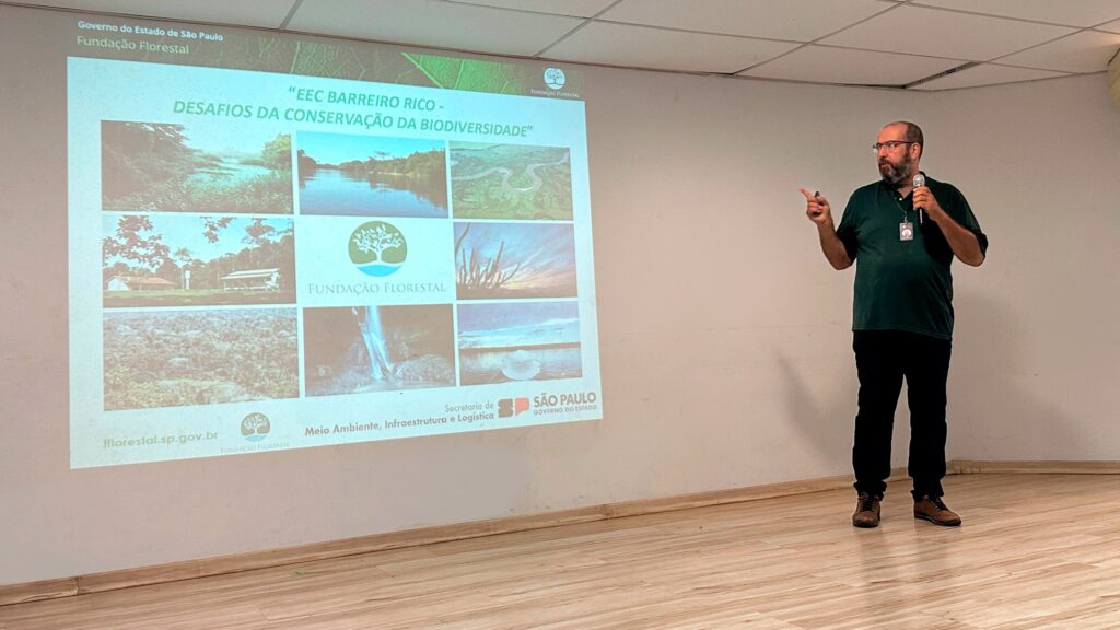 A imagem mostra uma apresentação de slides sobre os desafios da conservação da biodiversidade na Estação Ecológica do Barreiro Rico, localizada no Estado de São Paulo. O apresentador, um homem de meia-idade com óculos e barba, está usando uma camisa verde escura e calças pretas, e está gesticulando enquanto fala. Os slides mostram fotos da paisagem da região, incluindo rios, cachoeiras, vegetação e construções. A apresentação é organizada pela Fundação Florestal do Governo do Estado de São Paulo.