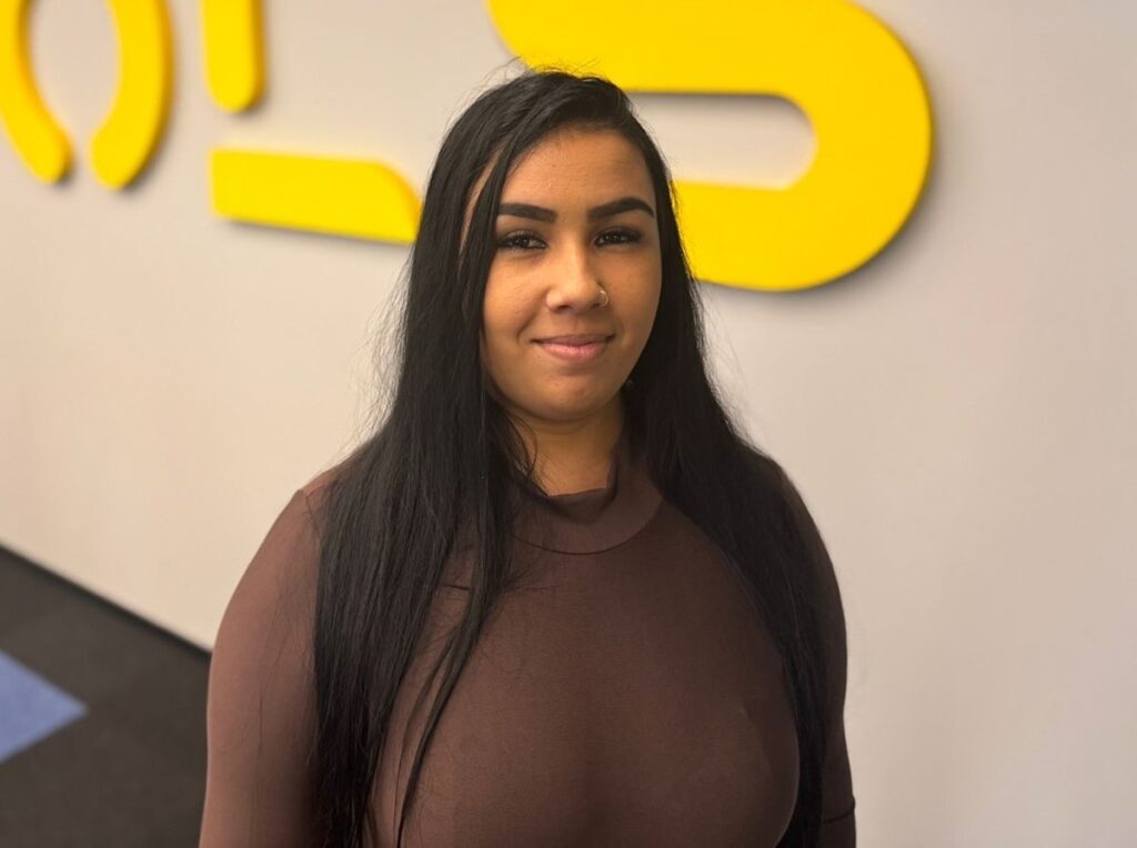 A imagem mostra uma jovem com cabelo longo e preto, vestindo uma blusa marrom escura de gola alta. Ela está parada na frente de uma parede com uma forma amarela vibrante na parede atrás dela. Ela parece estar sorrindo levemente para a câmera. A iluminação é suave e a imagem tem uma qualidade geral limpa e profissional.