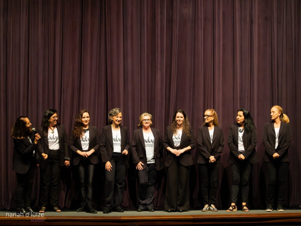 A imagem mostra dez mulheres em pé em um palco, alinhadas em duas fileiras. Elas estão vestindo roupas escuras, principalmente pretas, com blusas brancas por baixo dos casacos. As mulheres parecem estar em um evento formal, como uma premiação ou apresentação. O fundo é uma cortina de veludo marrom-escuro que cobre todo o palco. O ambiente é bem iluminado, e a fotografia é nítida e bem focada. Há um logotipo discreto no canto inferior esquerdo da imagem. As mulheres parecem ser adultas e têm aparências diversas. Algumas delas estão sorrindo para a câmera, enquanto outras mantêm expressões mais neutras. A atmosfera geral da imagem é formal e profissional.