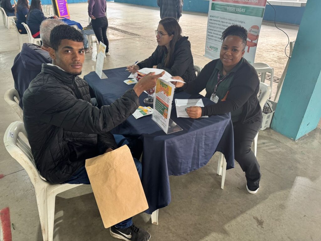 A imagem mostra uma cena em um evento de recrutamento. Há três pessoas sentadas em uma mesa, duas mulheres e um homem. Uma das mulheres parece ser uma recrutadora, pois está sentada atrás de uma mesa com documentos e folhetos. O homem parece ser um candidato a emprego, pois está olhando para a recrutadora e segurando um envelope pardo. A outra mulher também parece ser uma recrutadora. Ao fundo, há outras pessoas sentadas em cadeiras, possivelmente outros candidatos a emprego ou acompanhantes. Há um cartaz atrás das recrutadoras anunciando várias vagas e benefícios. O ambiente parece ser um local público, como uma praça de eventos ou um ginásio. A atmosfera parece tranquila e organizada.