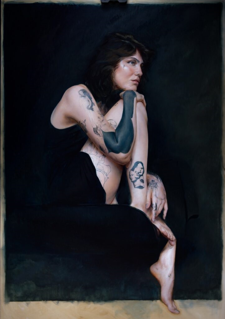A imagem é uma pintura a óleo de uma mulher sentada numa cadeira. Ela está vestida de preto e tem várias tatuagens no corpo. O fundo é escuro e a iluminação é suave, o que dá à pintura uma atmosfera sombria e misteriosa. A mulher parece pensativa e está virada para a direita, olhando para longe do espectador. As tatuagens são detalhadas e parecem realistas. O estilo da pintura é realista e a atenção aos detalhes é impressionante. A pintura evoca uma sensação de melancolia e introspecção.