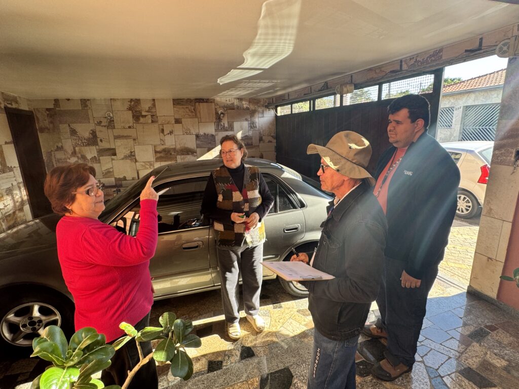 A imagem mostra quatro pessoas em uma garagem, aparentemente envolvidas em alguma espécie de discussão ou transação. Uma mulher de camisa vermelha está apontando para um carro cinza estacionado na garagem, enquanto uma outra mulher, vestindo um colete multicolorido, observa-a. Um homem de chapéu e um homem de jaqueta escura estão observando a conversa, com um deles segurando uma prancheta. O cenário é uma garagem com paredes de azulejos e uma porta de garagem aberta que revela um pouco do exterior. Há um carro adicional visível do lado de fora da garagem. A iluminação sugere que é dia, com a luz do sol entrando pela porta da garagem. A atmosfera parece ser informal, mas focada na interação entre as quatro pessoas. A imagem poderia ser interpretada como uma avaliação de um carro, uma discussão sobre uma compra, ou outra interação semelhante.