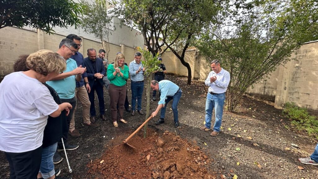 A imagem mostra um grupo de pessoas reunidas em torno de uma árvore sendo plantada pelo prefeito de Piracicaba, Helinho Zanatta,de camisa azul e calça jeans azul, segurando uma enxada e plantando uma árvore. Há pelo menos dez pessoas na foto, todas parecendo adultas. Elas estão vestidas com roupas casuais, e algumas estão aplaudindo. O cenário parece ser um pátio ou quintal, com árvores e um muro de concreto ao fundo. O chão é de terra. A atmosfera parece ser festiva e comunitária. A imagem sugere um evento de plantio de árvores ou uma atividade similar, possivelmente com um significado simbólico. A iluminação sugere que a foto foi tirada durante o dia.