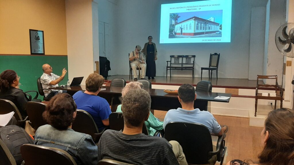 A imagem mostra uma apresentação em um auditório. No palco, um homem mais velho com bengala e uma mulher estão em pé, aparentemente apresentando algo. Na plateia, várias pessoas estão sentadas em cadeiras, prestando atenção. Há uma tela de projeção exibindo uma imagem de um prédio histórico e informações sobre o Museu Histórico e Pedagógico Prudente de Moraes em Piracicaba - SP. O auditório parece ser um espaço modesto, com cadeiras e uma mesa simples. A atmosfera parece ser formal e acadêmica, sugerindo uma palestra ou apresentação relacionada à história local. Há também um quadro na parede com uma foto e uma legenda. Um homem está fazendo gestos enquanto fala com a plateia. Um ventilador está visível no canto direito da imagem.