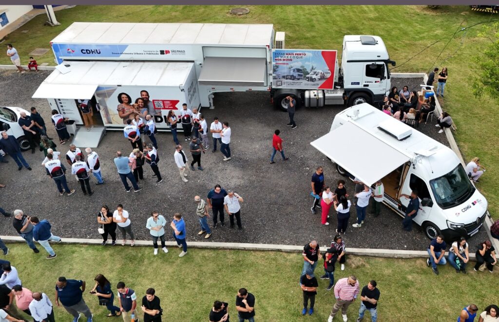 A imagem aérea mostra um evento ao ar livre com duas unidades móveis da CDHU (Companhia de Desenvolvimento Habitacional e Urbano) em destaque. Uma é um grande trailer com uma lona retrátil, e a outra é uma van menor com uma cobertura retrátil. Muitas pessoas estão reunidas em torno das unidades, algumas aparentemente interagindo com funcionários da CDHU, que usam coletes com o logotipo da companhia. Há também um grupo de pessoas sentadas em mesas sob uma árvore, e outras andando pelo gramado. A imagem sugere um evento de divulgação ou atendimento da CDHU, possivelmente em uma comunidade. Os veículos estão claramente marcados com a logomarca da CDHU e o logo do Estado de São Paulo. O cenário é um espaço aberto, com grama e uma área pavimentada.