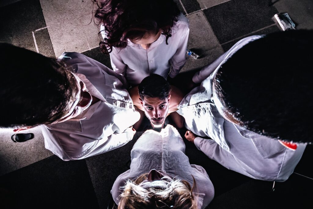 A imagem mostra cinco pessoas vestidas com camisas brancas, sentadas em círculo em torno de uma sexta pessoa, que é o foco central. O foco central é um homem sem camisa com o cabelo escuro e curto, com uma expressão séria no rosto. As outras quatro pessoas estão sentadas em torno dele, com suas cabeças inclinadas para baixo, olhando para ele. A pessoa no canto superior esquerdo parece ser uma mulher com cabelos escuros e compridos, enquanto as outras três pessoas parecem ser homens, com cabelos escuros e compridos. O fundo é escuro e o chão parece ser de ladrilhos. A iluminação é escura, mas o foco central está bem iluminado. A imagem tem uma atmosfera dramática e tensa.