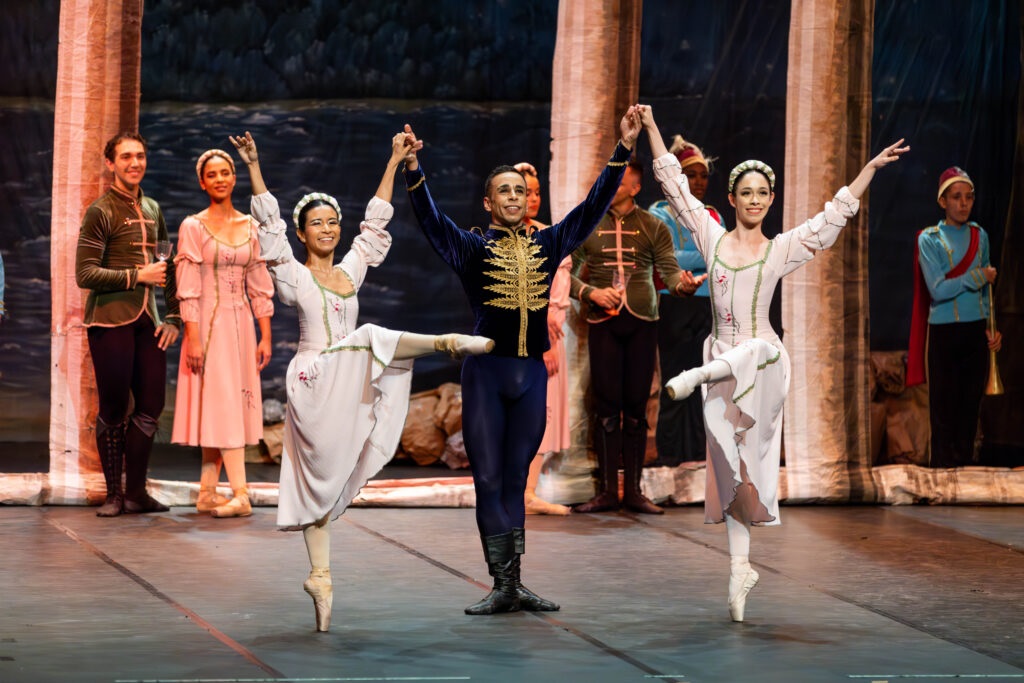 A imagem mostra um palco de teatro com bailarinos em trajes de época. No centro, um bailarino masculino está em pose com duas bailarinas femininas, cada uma com uma perna levantada em arabesque. Os bailarinos masculinos e femininos usam trajes elaborados, e os bailarinos femininos usam saias longas e brancas. No fundo, outros bailarinos podem ser vistos, bem como um cenário que parece ser um cenário de teatro. A iluminação é aconchegante e a atmosfera é elegante.