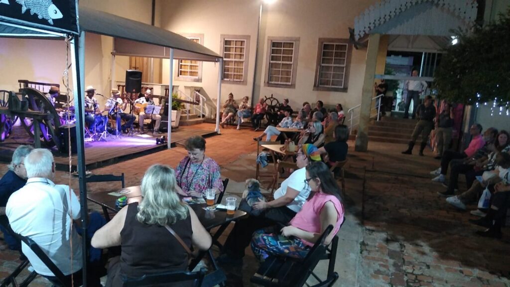 A imagem mostra uma cena noturna ao ar livre em um pátio de tijolos, onde um grupo de pessoas está assistindo a uma apresentação musical. Um pequeno palco está situado sob uma cobertura de lona, ​​e uma banda de quatro músicos está tocando instrumentos como guitarras, baixo e bateria. As luzes do palco iluminam os músicos, criando um contraste com o fundo mais escuro. A multidão está sentada em cadeiras de plástico, em mesas baixas e no chão, aparentemente relaxando e curtindo a música. As pessoas estão vestidas com roupas casuais, e há uma mistura de idades e gêneros presentes. Uma pessoa está com um cachorro pequeno. O fundo da imagem mostra o edifício onde o evento está acontecendo, que parece ser de arquitetura antiga, com paredes de tijolo claro e janelas grandes. Uma roda de madeira está visível perto do palco, o que sugere que o local pode ter algum significado histórico ou cultural. A atmosfera geral da imagem é descontraída e festiva, com uma sensação de comunidade e entretenimento ao ar livre. Há uma iluminação suave, que sugere uma atmosfera aconchegante e relaxante.