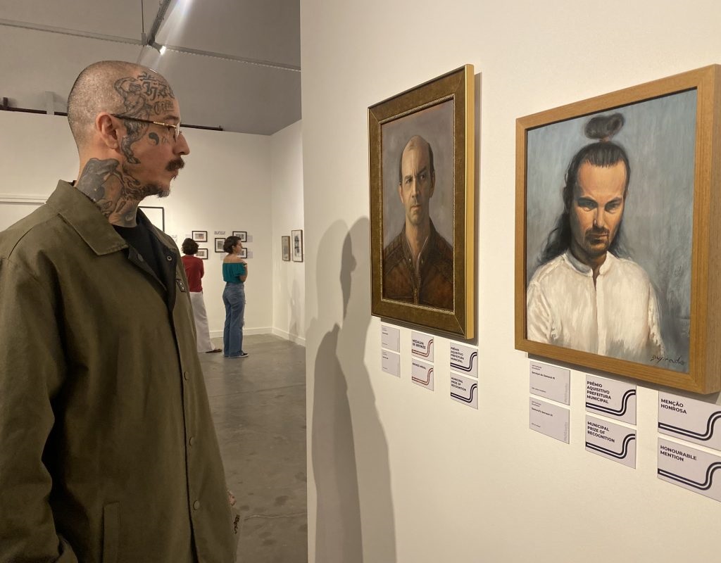 A imagem mostra um homem com tatuagens na cabeça e pescoço observando duas pinturas em uma galeria de arte. As pinturas são retratos, um de um homem careca e outro de um homem com cabelo longo preso em um coque. Há várias pessoas no fundo, incluindo duas mulheres que parecem estar olhando para outras obras de arte. As paredes são brancas e há cartões de reconhecimento municipal pendurados ao lado das pinturas. A atmosfera é calma e contemplativa.