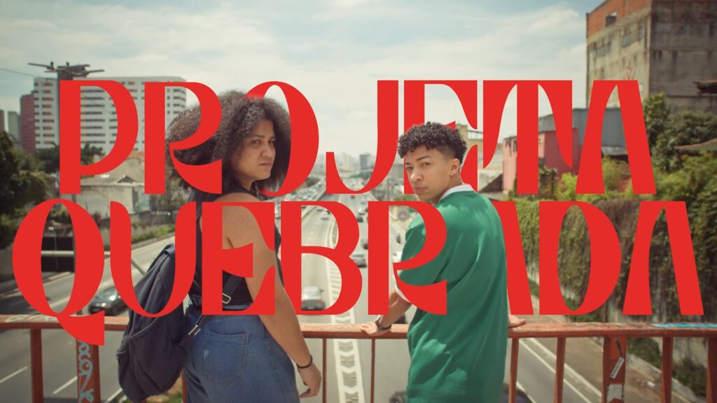A imagem mostra dois jovens adultos, uma mulher e um homem, de pé em um viaduto com uma rodovia movimentada ao fundo. Ambos estão voltados para a câmera, mas ligeiramente para o lado, criando uma composição dinâmica. A mulher está à esquerda, vestindo uma camiseta preta e jeans, com uma mochila nas costas. O homem está à direita, vestindo uma camisa verde escura de manga comprida. Ambos parecem estar em seus vinte anos e têm expressões sérias. O título "PROJETA QUEBRADA" está sobreposto à imagem em letras grandes e vermelhas, criando um contraste marcante com as cores mais suaves do fundo. As letras são estilizadas, com uma aparência moderna e ousada. O fundo é uma paisagem urbana brasileira, com edifícios e uma rodovia visíveis. A imagem é bem iluminada, com um tom geral vibrante. A composição e a arte visual sugerem um enquadramento cinematográfico, talvez um pôster de filme ou uma captura de tela de um filme. A imagem como um todo comunica uma sensação de juventude, urbanidade e talvez uma mensagem social implícita, dada a localização e o título.