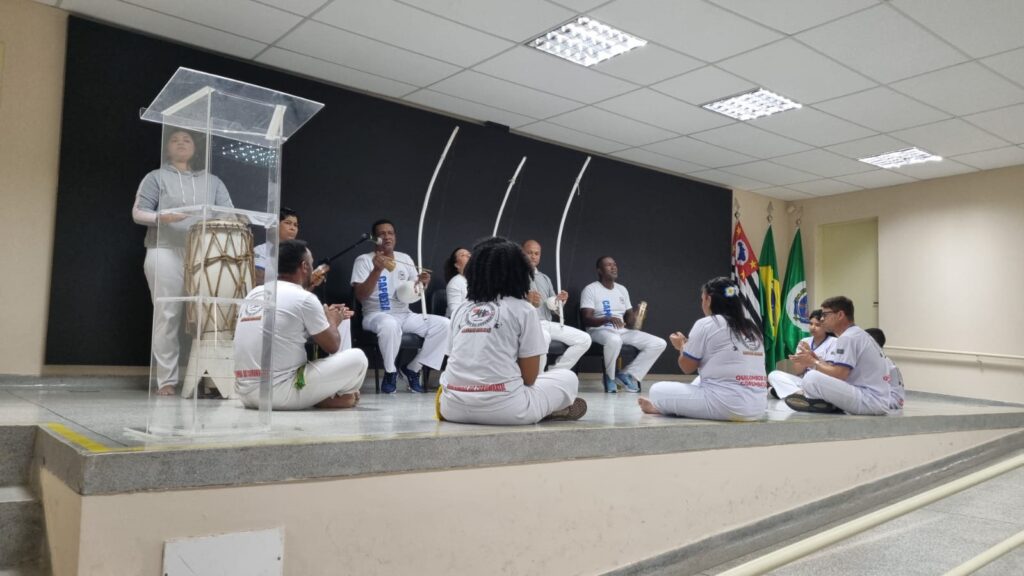 A imagem mostra um grupo de pessoas sentadas em círculo no palco, tocando instrumentos musicais tradicionais brasileiros. Há um berimbau, um atabaque e outros instrumentos. As pessoas estão vestindo roupas brancas e parecem estar praticando capoeira. Uma mulher está de pé atrás de um púlpito, possivelmente dando uma apresentação ou instrução. No fundo, há bandeiras brasileiras. A cena parece ser uma apresentação ou aula de capoeira. O ambiente é um espaço interno, possivelmente uma escola ou centro comunitário. A iluminação é artificial, provinda de luminárias no teto. A imagem é nítida e bem iluminada.