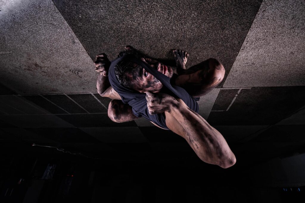 A imagem mostra um homem enrolado em um tecido com um olhar fixo para cima. Ele está agachado em um piso liso nas cores clara e escuro.