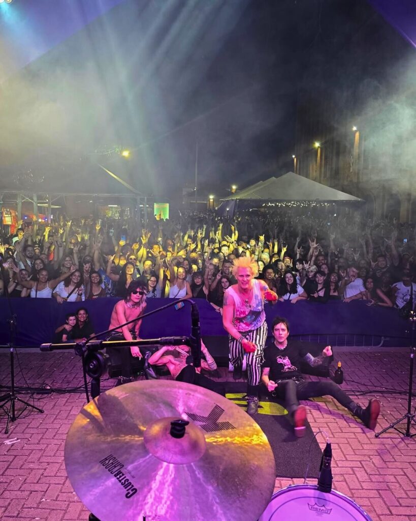 A imagem mostra uma banda se apresentando ao ar livre para uma grande multidão. O palco está situado em uma área pavimentada, e a multidão está atrás de uma barreira baixa. O palco é relativamente simples, com um conjunto de bateria e alguns microfones visíveis. A banda parece ser um grupo de rock, com os membros vestindo roupas coloridas e ousadas. A multidão está animada e engajada, com muitas pessoas levantando as mãos e cantando. A iluminação é escura, com holofotes brilhantes no palco, criando uma atmosfera energética e vibrante. O fundo mostra prédios e outras estruturas de uma cidade, conferindo um contexto urbano ao evento. A atmosfera geral da imagem sugere um concerto ao vivo emocionante e animado.