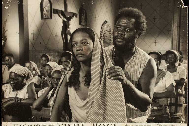 A imagem é um close de uma cena de filme em preto e branco, aparentemente de um filme brasileiro chamado "Sinhá Moça". Mostra uma mulher negra com um lenço na cabeça, com uma mancha de sangue no rosto, sentada ao lado de um homem negro de barba. Vários outros indivíduos negros estão sentados ao fundo, em uma igreja ou local de culto. A iluminação é escura, com destaque para os personagens principais. A atmosfera é tensa e dramática. A imagem sugere uma narrativa envolvendo sofrimento e possivelmente injustiça. O estilo fotográfico é típico de filmes antigos, com granulação e contraste acentuados. A imagem é uma publicidade do filme, com o título e os créditos visíveis na parte inferior.