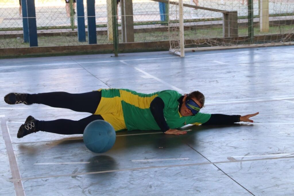 A imagem mostra uma jogadora de goalball feminina deitada de bruços no chão de uma quadra de esportes. Ela está usando uma venda nos olhos e uma camisa verde e amarela, e está segurando uma bola de goalball azul. A quadra parece estar dentro de um ginásio, e há uma cerca de arame atrás dela. A jogadora parece estar se preparando para jogar ou esperando sua vez de jogar.