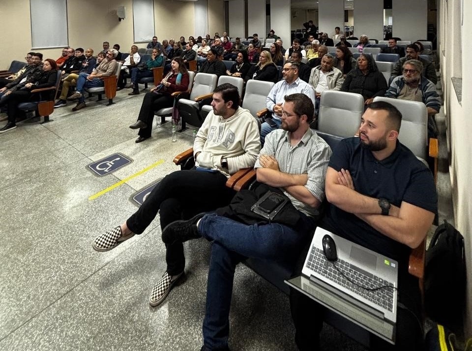 A imagem mostra uma sala de conferências ou auditório com muitas pessoas sentadas em cadeiras dispostas em fileiras. A sala é bem iluminada e as pessoas parecem estar assistindo a uma apresentação ou palestra. Há uma mistura de homens e mulheres, e a maioria deles parece estar vestindo roupas casuais. Algumas pessoas estão sentadas sozinhas, enquanto outras estão sentadas em grupos. Há também algumas cadeiras vazias na sala. No chão, há marcações que parecem indicar locais para pessoas com deficiência. No geral, a imagem sugere um evento formal ou semi-formal, como uma reunião, uma conferência ou uma apresentação.