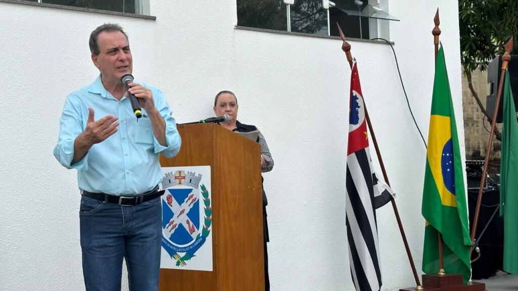 A imagem mostra o prefeito de Piracicaba, Helinho Zanatta, de camisa azul clara e jeans, falando em um microfone em um evento ao ar livre. Ele está parado atrás de um pódio de madeira com o brasão de uma cidade ou região. Ao fundo, uma mulher de vestido escuro escuta atentamente. Duas bandeiras estão em cada lado do pódio: uma bandeira brasileira e outra que parece ser uma bandeira estadual ou municipal. Ao fundo, há um prédio branco e alguma vegetação. A cena parece ser um evento formal ou cerimonial público. A iluminação é natural e o ambiente parece ser tranquilo e organizado. O homem parece estar fazendo um discurso ou apresentação. A mulher parece estar participando do evento como público ou convidada.