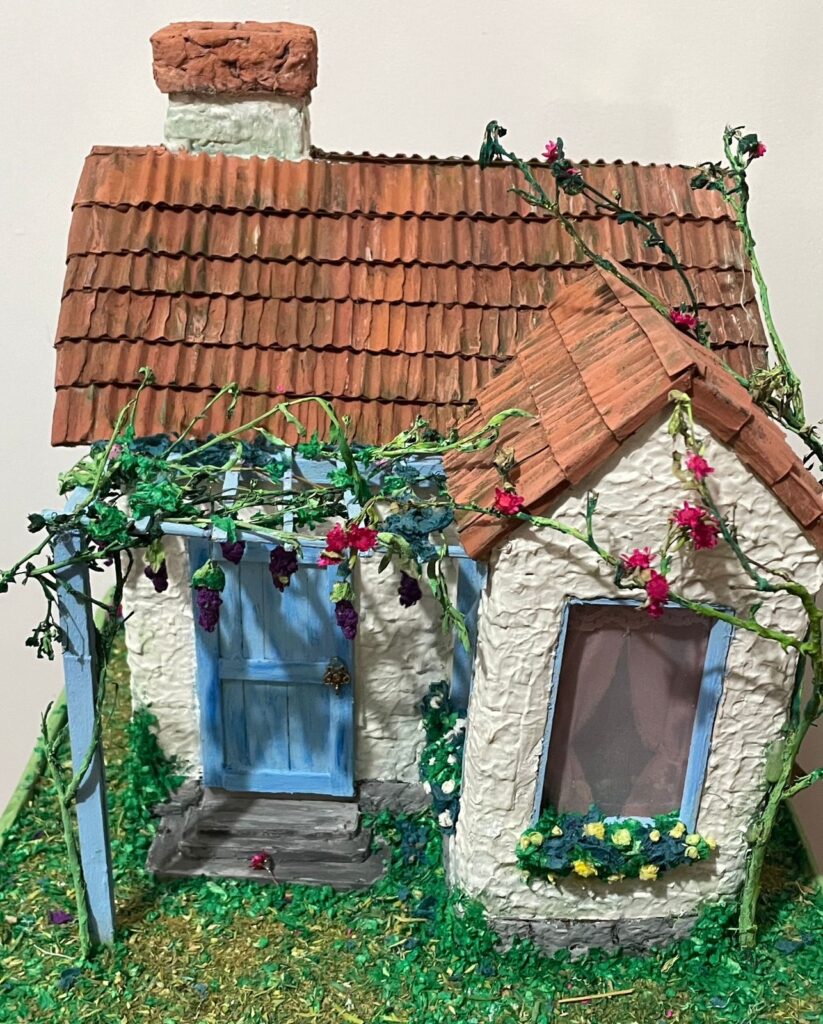 A imagem mostra uma maquete detalhada de uma pequena casa de campo. A casa é de estilo mediterrânico, com telhado de telha vermelha e paredes de estuque branco texturizado. Há uma porta azul e uma janela com um vaso de flores embaixo. Videiras com uvas e flores crescem em uma pérgola sobre a porta da frente e ao longo das paredes. A casa está sentada em um pequeno jardim com grama e outras plantas. O nível de detalhe é impressionante, com texturas realistas e cores vibrantes que dão vida à cena.