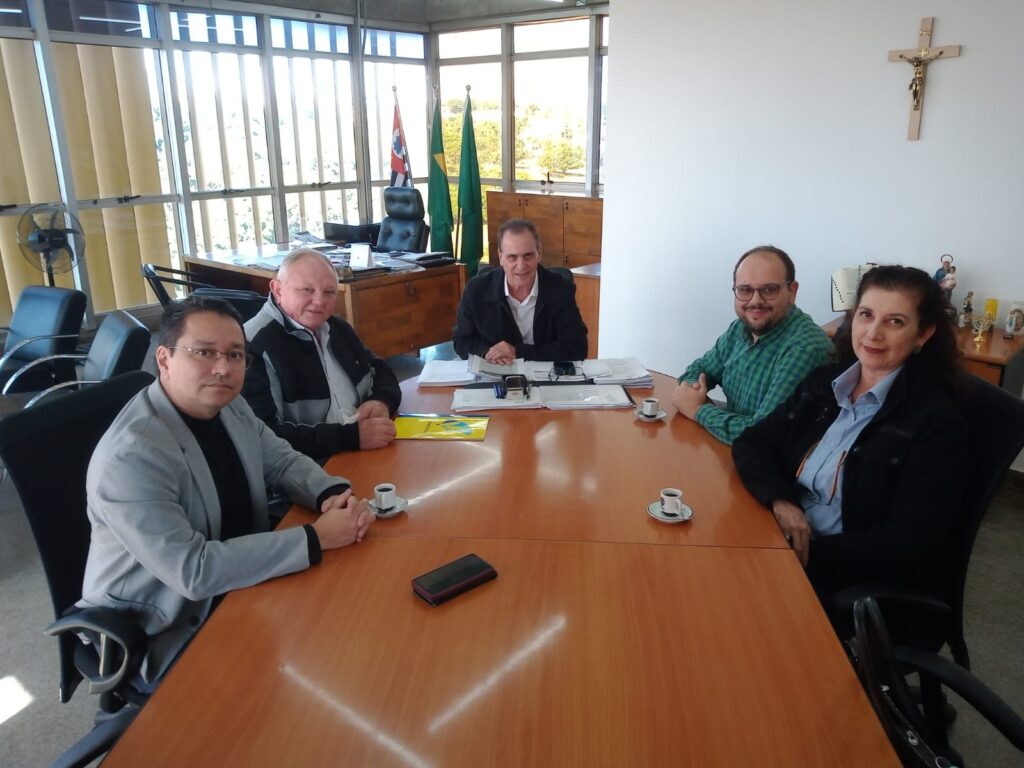 Cinco pessoas estão reunidas ao redor de uma mesa de reuniões, sorrindo para a câmera em um ambiente formal e bem iluminado. No fundo, aparecem bandeiras oficiais, um crucifixo na parede e objetos decorativos sobre um móvel. Pequenas xícaras de café estão dispostas sobre a mesa, indicando um encontro cordial e institucional.
