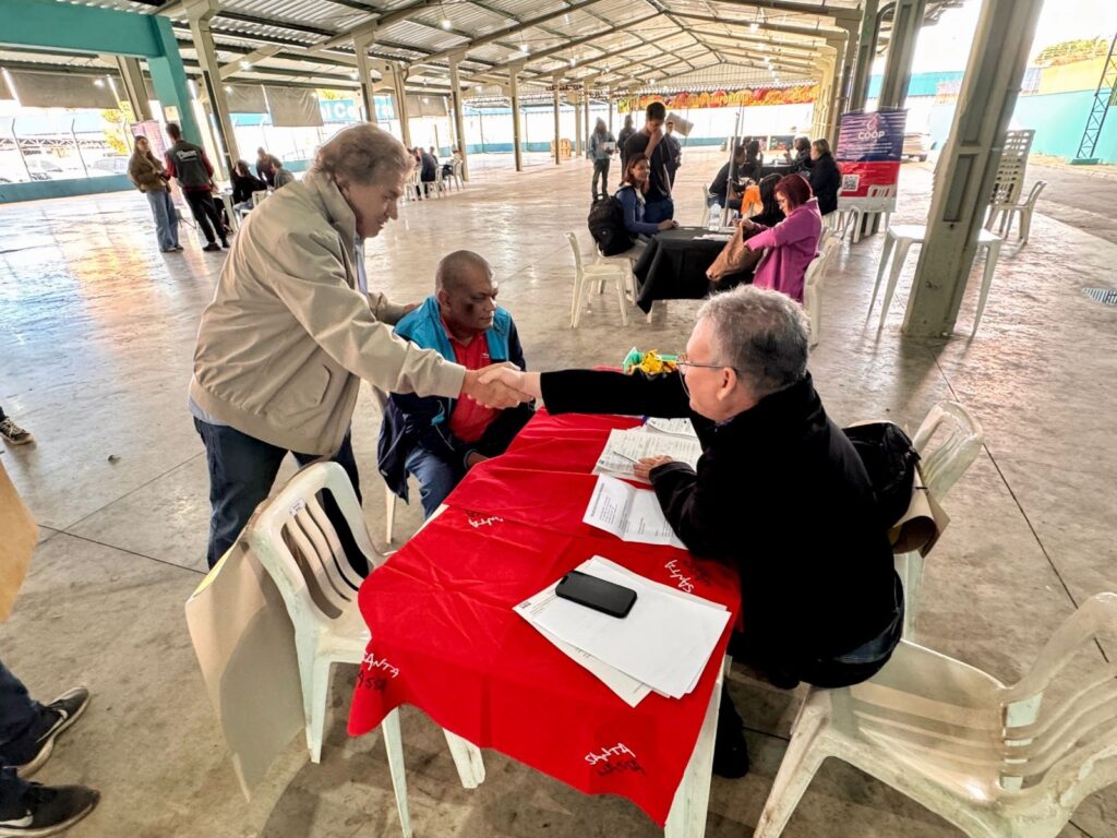A imagem mostra uma cena em um grande galpão aberto, aparentemente um local de atendimento ao público. Há várias pessoas sentadas em mesas, algumas interagindo com atendentes. O chão é de concreto polido, e a estrutura do galpão é de metal aparente e telhado translúcido. No centro da imagem, o foco principal é em três pessoas: um homem mais velho de jaqueta bege cumprimentando um homem mais jovem de camisa vermelha, enquanto um terceiro homem, sentado à mesa, parece estar atendendo-os. Há documentos e um celular sobre a mesa coberta com uma toalha vermelha. Ao redor, outras pessoas aguardam ou são atendidas em mesas similares. Muitas cadeiras de plástico branco estão espalhadas pelo local. A atmosfera parece ser calma e organizada, apesar da quantidade de pessoas presentes. A iluminação é natural, vinda do telhado translúcido do galpão. O ambiente sugere um evento comunitário, uma feira de serviços ou um processo de atendimento público em uma área aberta.