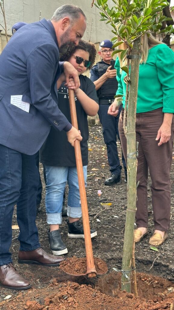 A imagem mostra um grupo de pessoas plantando uma árvore. Uma criança está usando óculos de sol e ajudando a cavar um buraco com uma pá. Um homem de terno está ajoelhado, também ajudando a plantar a árvore. Uma mulher vestida de verde está ao lado da árvore, observando. Há outro homem uniformizado atrás deles, que parece ser um policial. O cenário parece ser uma rua ou um parque. O solo é marrom e há algumas folhas no chão. A atmosfera é calma e pacífica.