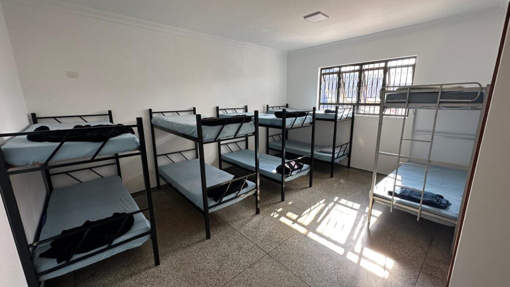 A imagem mostra um quarto com várias beliches. As paredes são brancas e o chão é de granito. Há uma janela grande na parede do fundo que deixa entrar bastante luz solar. As beliches são de metal preto e têm colchões azuis claros. Cada beliche tem um cobertor preto dobrado em cima do colchão. Há também uma beliche de metal prateado no canto direito da imagem. O quarto parece ser um dormitório ou um alojamento.