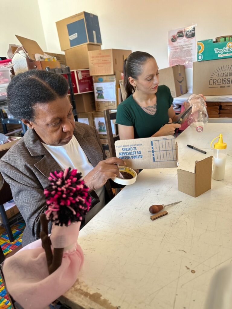 A imagem mostra duas mulheres sentadas em uma mesa, envolvidas em uma atividade artesanal. A mulher mais velha, à esquerda, está usando uma jaqueta marrom e está envolvida na montagem de uma boneca de pano. Ela está usando fita adesiva para fixar algo. A mulher mais jovem, à direita, está usando uma camiseta verde e está cortando uma garrafa de plástico com uma tesoura. Ambas as mulheres parecem estar trabalhando em um projeto de artesanato com materiais reciclados, como caixas de papelão e garrafas de plástico. Ao fundo, há muitas caixas de papelão empilhadas, sugerindo um ambiente de armazenamento ou oficina. A mesa está coberta de vários materiais de artesanato, incluindo tesoura, fita adesiva, garrafa de plástico cortada, caixas de papelão e uma pequena boneca de pano. Há também uma pequena garrafa de cola na mesa. A atmosfera geral da imagem é calma e focada, sugerindo um trabalho colaborativo e criativo.
