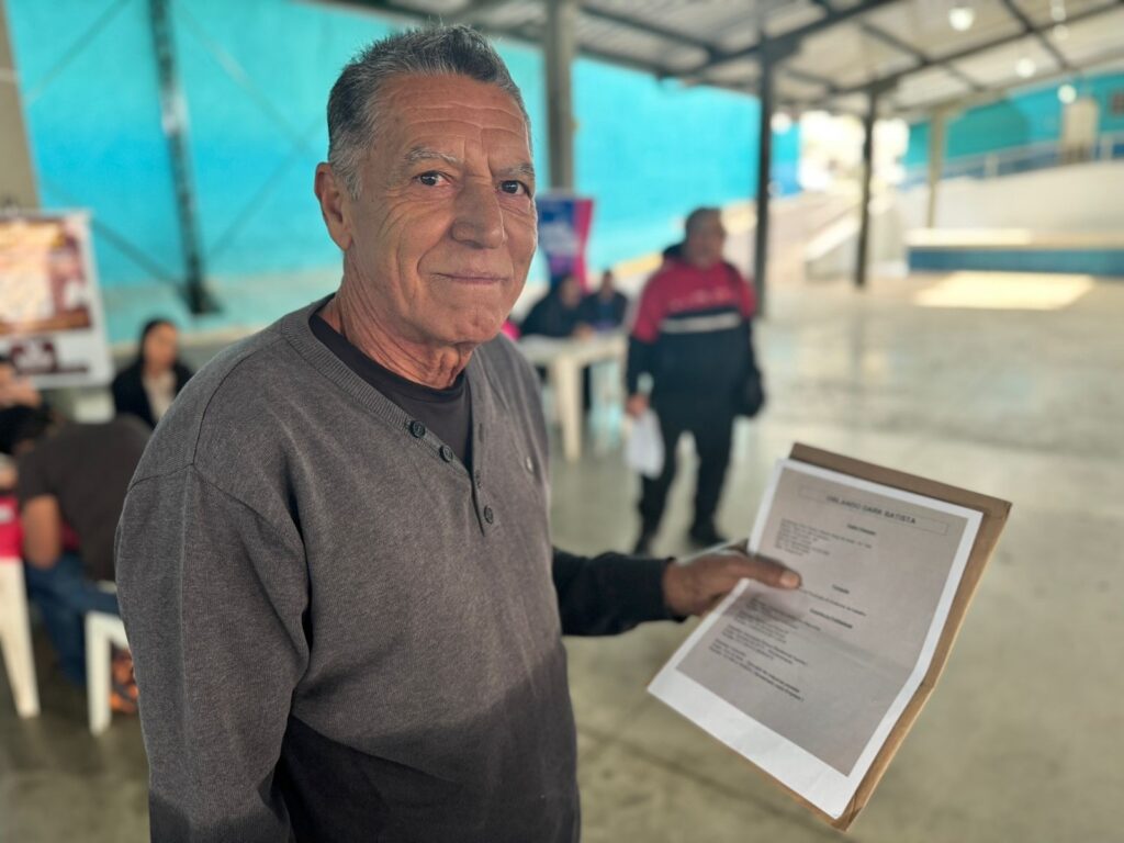 A imagem mostra um homem de meia-idade, de pele morena e cabelos grisalhos penteados para trás, vestindo um suéter cinza escuro de gola alta com botões. Ele está sorrindo levemente para a câmera e segurando um documento em suas mãos. O documento parece ser um formulário ou algum tipo de papelada oficial, com texto impresso nele. O fundo é desfocado, mas mostra um ambiente interno, possivelmente um espaço público ou comunitário, com outras pessoas presentes ao fundo, algumas sentadas em mesas. As cores do ambiente são neutras e o clima parece ser calmo e tranquilo. O foco principal da imagem é o homem e o documento que ele está segurando, sugerindo que ele possa estar envolvido em algum tipo de processo administrativo ou burocrático.