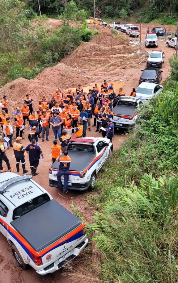 A imagem mostra uma cena ao ar livre, aparentemente em uma área rural ou de acesso difícil, com um grupo numeroso de pessoas reunidas. Elas parecem estar uniformizadas, possivelmente pertencentes a alguma organização de defesa civil ou resgate, considerando a presença de veículos com a inscrição "Defesa Civil". Há vários carros e caminhonetes estacionados ao longo de uma estrada de terra batida, alguns deles com sinalização oficial. A estrada apresenta marcas de erosão ou passagem de veículos, e o terreno ao redor é irregular, com terra exposta e vegetação esparsa. A cena sugere uma situação de emergência ou resposta a algum evento, como um desastre natural ou acidente, devido à concentração de pessoas e veículos de emergência. A vegetação ao redor indica um clima possivelmente tropical ou subtropical. A iluminação sugere que a foto foi tirada durante o dia.