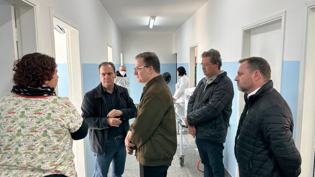 A imagem mostra um corredor de um hospital ou instalação médica, com várias pessoas paradas e conversando. O corredor é longo e estreito, com paredes brancas e portas azuis claras em cada lado. O chão é de um material claro, possivelmente ladrilhos. Há sete pessoas na imagem. Uma mulher com cabelos castanhos encaracolados está de costas para a câmera, vestindo uma camisa com um padrão de crocodilos. Ela está conversando com um homem de meia-idade com cabelos escuros, vestindo uma jaqueta escura. Há outros quatro homens e uma mulher em pé perto deles. Os homens estão vestindo jaquetas e a mulher está vestindo um jaleco branco. No fundo, há um carrinho de transporte médico. A iluminação é suave e uniforme, sugerindo que a imagem foi tirada dentro de casa. A atmosfera geral é calma e profissional.