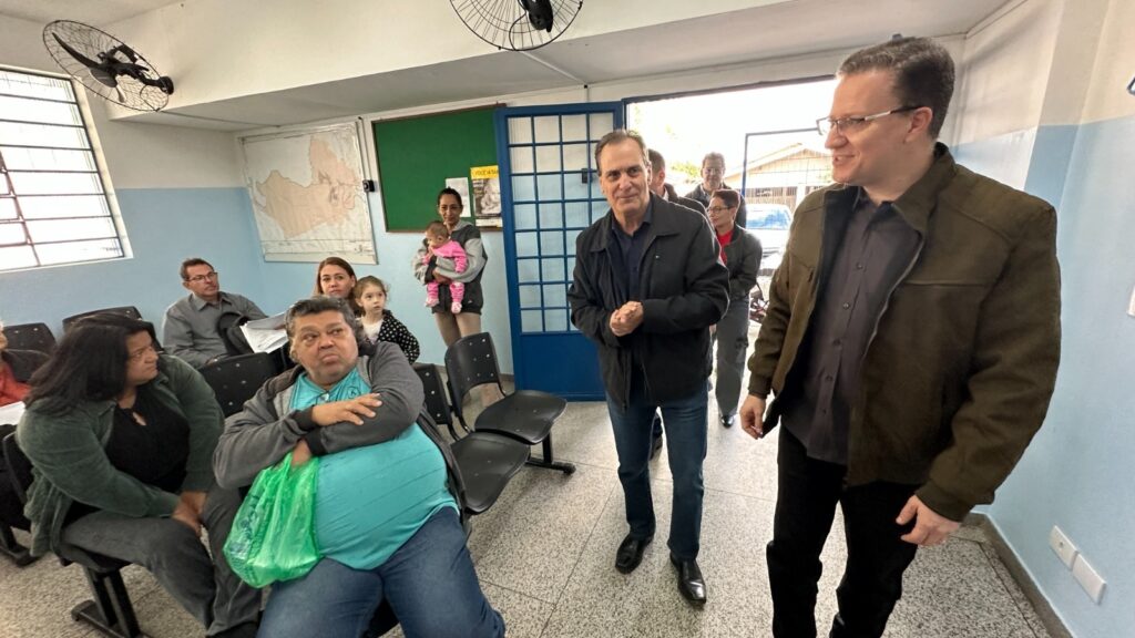 A imagem mostra uma sala de espera, possivelmente em um consultório médico ou centro comunitário. Há várias pessoas sentadas em cadeiras de plástico, algumas conversando entre si. Uma mulher com um bebê nos braços está de pé perto de uma parede. Um homem de terno escuro está de pé no centro da sala, parecendo estar em uma posição de autoridade ou liderança. Há um mapa na parede e alguns ventiladores de teto. A atmosfera geral parece calma e organizada. As pessoas na imagem parecem estar esperando por algo ou participando de uma reunião informal. A iluminação é suave, e as cores são predominantemente neutras, com tons de azul claro nas paredes e cores sóbrias nas roupas das pessoas. A imagem sugere um ambiente público ou semi-público, onde as pessoas estão reunidas para um propósito comum.