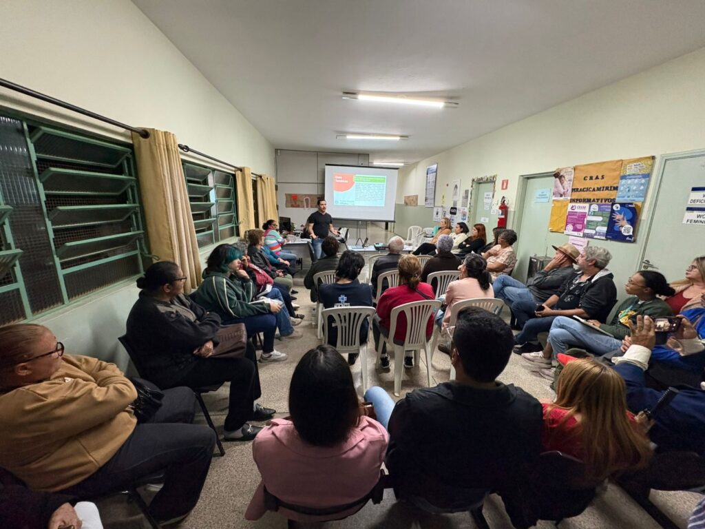 A imagem mostra uma sala de espera ou sala de aula lotada de pessoas sentadas em cadeiras de plástico brancas. Uma pessoa está dando uma apresentação na frente de uma tela de projeção, com slides visíveis. A sala é simples, com paredes de cor creme e janelas com cortinas bege. Há alguns cartazes nas paredes. As pessoas na sala parecem estar envolvidas na apresentação e estão prestando atenção ao palestrante. A imagem parece ter sido tirada de um ângulo baixo, dando uma perspectiva única da sala e das pessoas. Há uma variedade de pessoas na sala, com diferentes idades e gêneros. A atmosfera geral da imagem é calma e atenciosa.