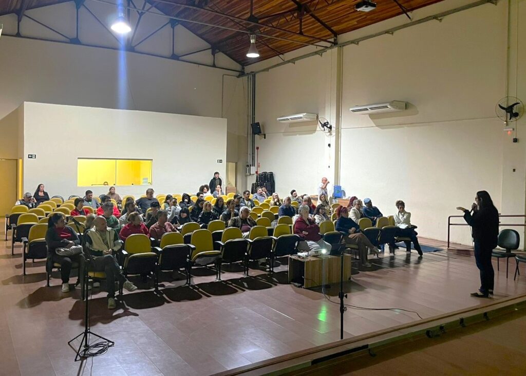 A imagem mostra uma sala de conferências ou auditório com muitas pessoas sentadas em cadeiras amarelas. Uma mulher está de pé no palco, falando ao microfone, aparentemente dirigindo-se ao público. A sala é espaçosa, com paredes claras e um teto alto com vigas de madeira expostas. Há alguns aparelhos de ar condicionado na parede e um ventilador de teto. A iluminação é moderada, com uma luz mais brilhante vindo do teto. A atmosfera parece ser formal e atenta, sugerindo uma apresentação ou palestra.