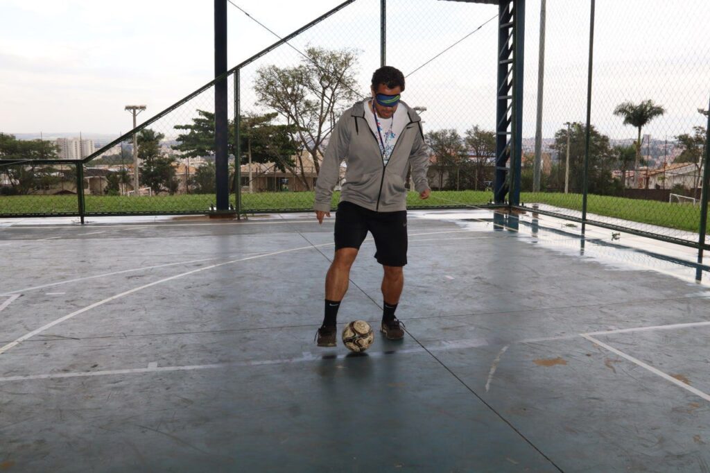 A imagem mostra um homem de meia-idade com uma máscara nos olhos, praticando futebol numa quadra ao ar livre. Ele está usando uma jaqueta cinza, shorts pretos e meias pretas. O homem está se concentrando na bola, que está no chão entre seus pés. A quadra é de concreto, com linhas brancas pintadas para marcar o campo de jogo. Há uma cerca de metal ao redor da quadra, e um fundo arborizado é visível além dela. O tempo parece nublado, e há alguma umidade no chão. A imagem sugere um exercício de coordenação motora ou um desafio para pessoas com deficiência visual. A expressão do homem é séria e concentrada, indicando sua dedicação à atividade.