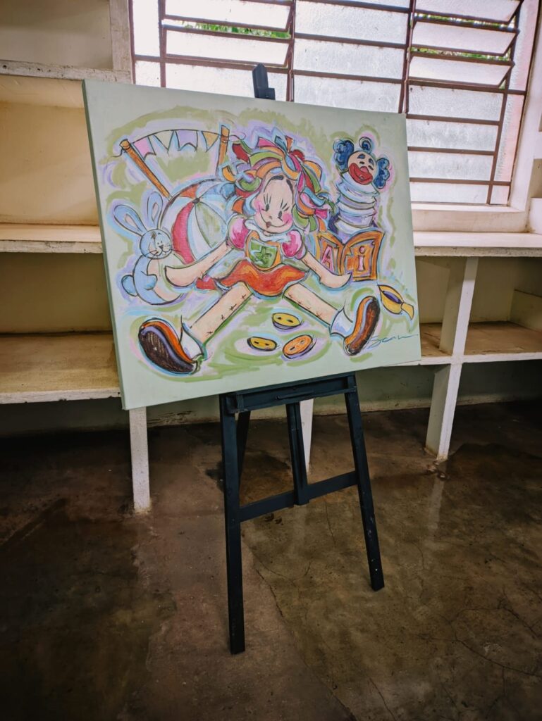 A imagem mostra uma tela apoiada em um cavalete. A tela apresenta uma pintura colorida e vibrante, aparentemente infantil, com uma boneca de pano como personagem principal, cercada por outros elementos como um coelho, uma bola e blocos de letras. A paleta de cores é alegre e pastel, com tons de verde, rosa, laranja e azul predominando. A pintura parece ter sido feita com pinceladas soltas e expressivas, conferindo-lhe um aspecto informal e espontâneo. A tela está posicionada em um ambiente interno, possivelmente um ateliê ou um espaço de trabalho artístico. O chão é de concreto, com aspecto um pouco irregular e sujo, e há um pequeno vazamento de água visível no chão. O cavalete é simples e de cor escura, contrastando com a luminosidade da pintura. A composição da imagem sugere uma cena tranquila e criativa.