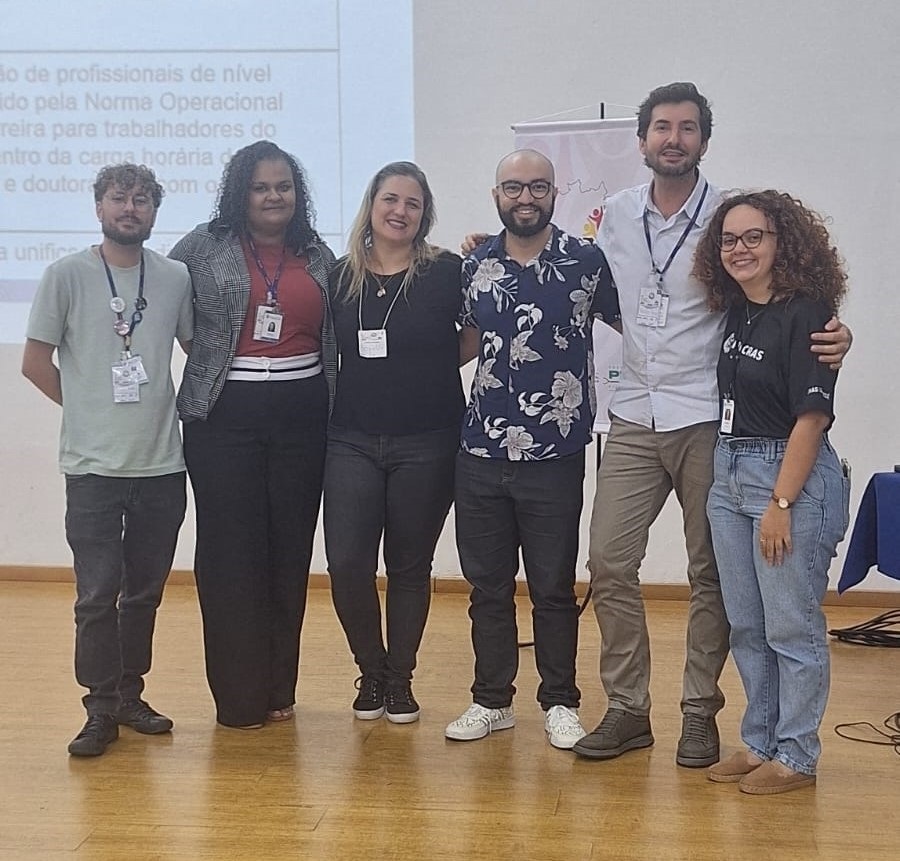 A imagem mostra seis indivíduos, cinco mulheres e um homem, em pé lado a lado, sorrindo para a câmera. Eles estão dispostos em duas fileiras, com três pessoas na fileira da frente e três na fileira de trás. Atrás deles, há uma apresentação em um projetor e um painel. As roupas são casuais, mas profissionais. As mulheres usam calças e blusas, enquanto o homem usa uma camisa de manga comprida e calças. Eles parecem ser um grupo de colegas de trabalho ou participantes de um evento. Há um clima amigável e colaborativo na foto.