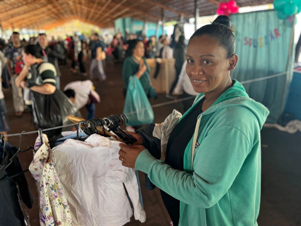 A imagem mostra uma mulher de pele escura, com o cabelo preso em um coque, sorrindo para a câmera. Ela está vestindo um casaco verde-água com capuz e parece estar em uma feira ou mercado ao ar livre. Ela está segurando algumas peças de roupa em cabides, que estão pendurados em uma corda. O fundo está bastante movimentado, com muitas outras pessoas que parecem estar comprando ou vendendo mercadorias. O ambiente parece ser uma área coberta, talvez uma estrutura de madeira temporária ou um galpão. Há outras pessoas ao fundo, desfocadas, mas visíveis. A iluminação parece ser natural, provavelmente luz do dia. A mulher parece estar focada em suas mercadorias, mas também está engajada com o ambiente ao seu redor. A atmosfera geral sugere um evento comunitário ou um mercado de pulgas informal. A mulher exibe uma expressão amigável e acessível.