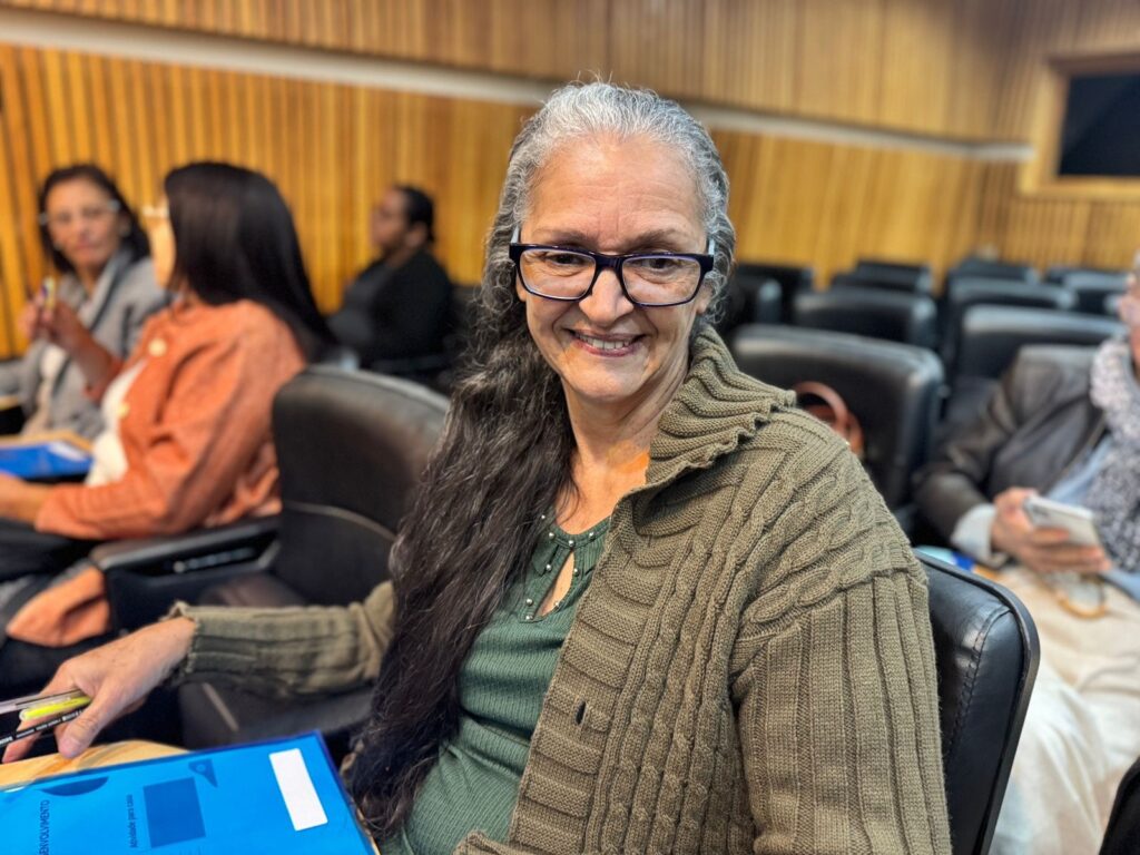 A imagem mostra uma mulher de meia-idade com cabelos grisalhos longos e óculos, sentada em uma cadeira em uma sala de palestras ou auditório. Ela está sorrindo para a câmera e parece confortável e relaxada. Ela veste um casaco de tricô verde-oliva sobre uma blusa verde escura. A mulher segura uma pasta azul clara em seu colo. Ao fundo, outras pessoas estão sentadas em cadeiras, levemente desfocadas, indicando que o foco principal da imagem é a mulher em primeiro plano. As pessoas ao fundo parecem estar em uma reunião ou apresentação. A sala é bem iluminada e tem paredes de madeira. Os assentos são estofados e escuros. A expressão facial da mulher é amigável e acolhedora, contribuindo para uma atmosfera geral de calma e engajamento. A imagem parece ter sido tirada em um ambiente profissional ou acadêmico. A qualidade da imagem é boa, com boa resolução e cores vibrantes.