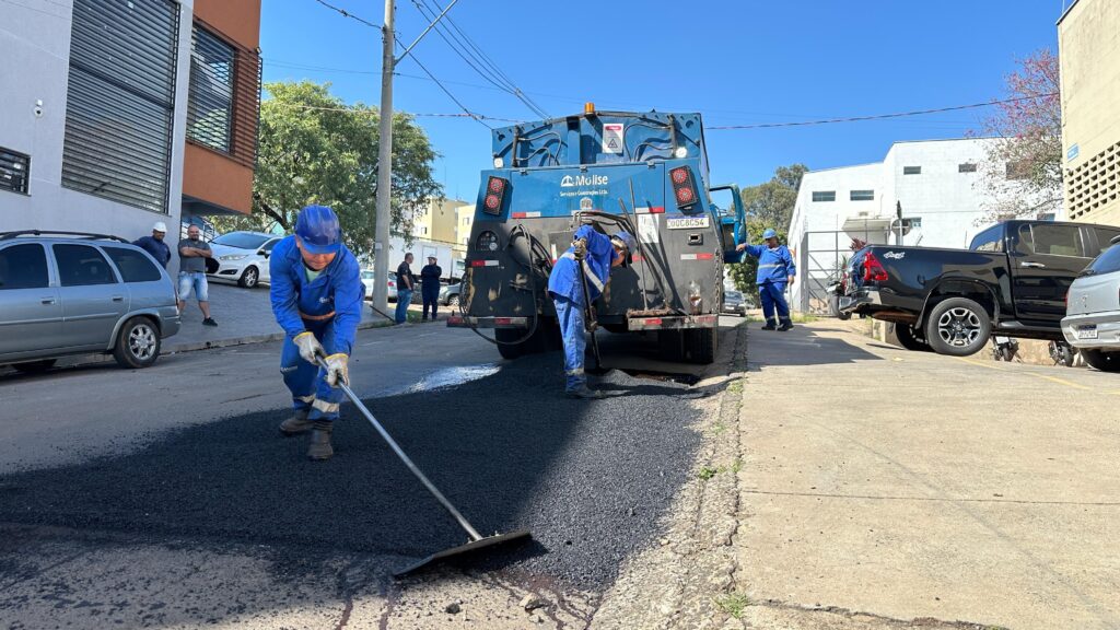A imagem mostra uma equipe de trabalhadores realizando reparos em uma rua. Um caminhão de serviço, azul e com a marca "Mulise", está estacionado no centro da cena, com trabalhadores usando roupas de trabalho azuis ao redor. Eles parecem estar aplicando asfalto novo em um buraco ou seção danificada da rua. Alguns trabalhadores usam pás ou ferramentas semelhantes para espalhar e nivelar o asfalto. Há alguns carros estacionados ao fundo, e algumas pessoas observando a cena a uma distância segura. O cenário parece ser uma rua residencial ou comercial, com edifícios residenciais ao fundo. O dia está ensolarado.