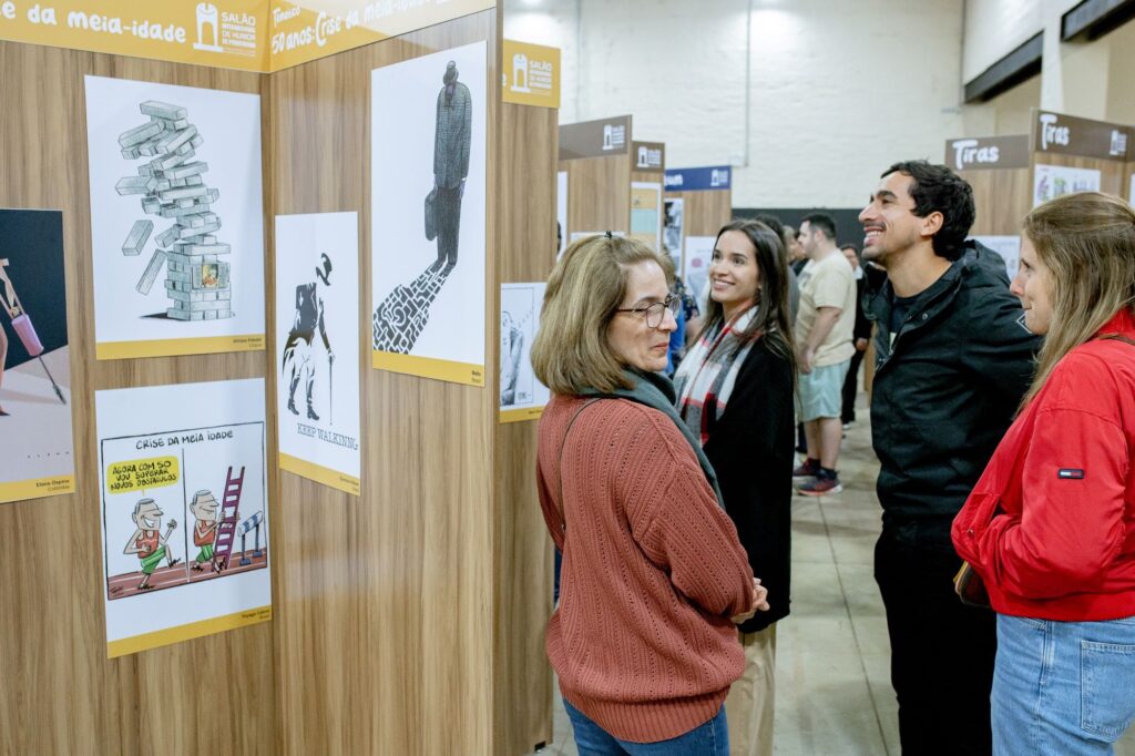 A imagem mostra uma exibição de arte em uma galeria ou espaço de evento. As paredes são de madeira clara, e várias placas de exibição apresentam desenhos de humor, muitos deles com temas relacionados à meia-idade. Os desenhos variam em estilo, alguns são cartuns enquanto outros são mais minimalistas. As legendas são visíveis em português, indicando que a exibição é provavelmente em um país de língua portuguesa. Pessoas estão presentes na galeria, observando as obras de arte. Há um grupo de mulheres conversando perto de uma das placas de exibição, e um homem está conversando com uma mulher perto de outra placa. As pessoas parecem estar envolvidas e interessadas na arte. O ambiente parece casual e acolhedor. A imagem é bem iluminada e nítida, permitindo que os detalhes dos desenhos e das expressões faciais das pessoas sejam facilmente observados. A fotografia é bem composta, com um bom equilíbrio entre as obras de arte e as pessoas presentes. A atmosfera geral é de um evento cultural positivo e envolvente.