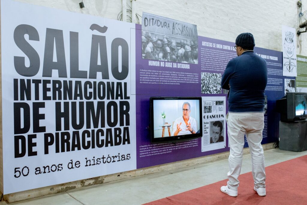A imagem mostra uma exposição comemorativa de 50 anos do Salão Internacional de Humor de Piracicaba. A exposição inclui painéis com textos e imagens que contextualizam o humor brasileiro durante a ditadura militar, destacando o papel dos artistas em resistir à censura e opressão através da sátira. Há também uma tela exibindo um vídeo, possivelmente com entrevistas ou documentário sobre o tema. Um homem de meia-idade observa atentamente os painéis. A atmosfera é sóbria, refletindo a seriedade do tema abordado, mas ao mesmo tempo vibrante pela riqueza de imagens e informações.