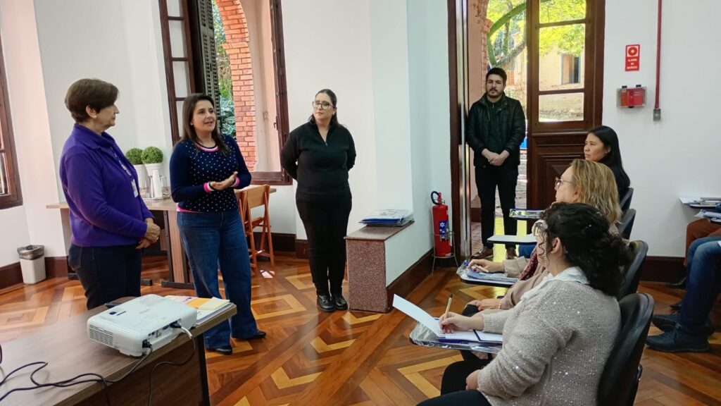 A imagem mostra uma reunião em uma sala. Há várias mulheres sentadas em mesas e cadeiras, algumas em pé. As mulheres estão vestidas com roupas formais e parecem estar participando de uma apresentação ou discussão. Há um projetor e um computador na mesa. A sala parece ser um escritório ou sala de aula. A atmosfera é profissional e calma. Há também um homem de pé perto da porta. No geral, a imagem sugere uma reunião de trabalho ou acadêmica.
