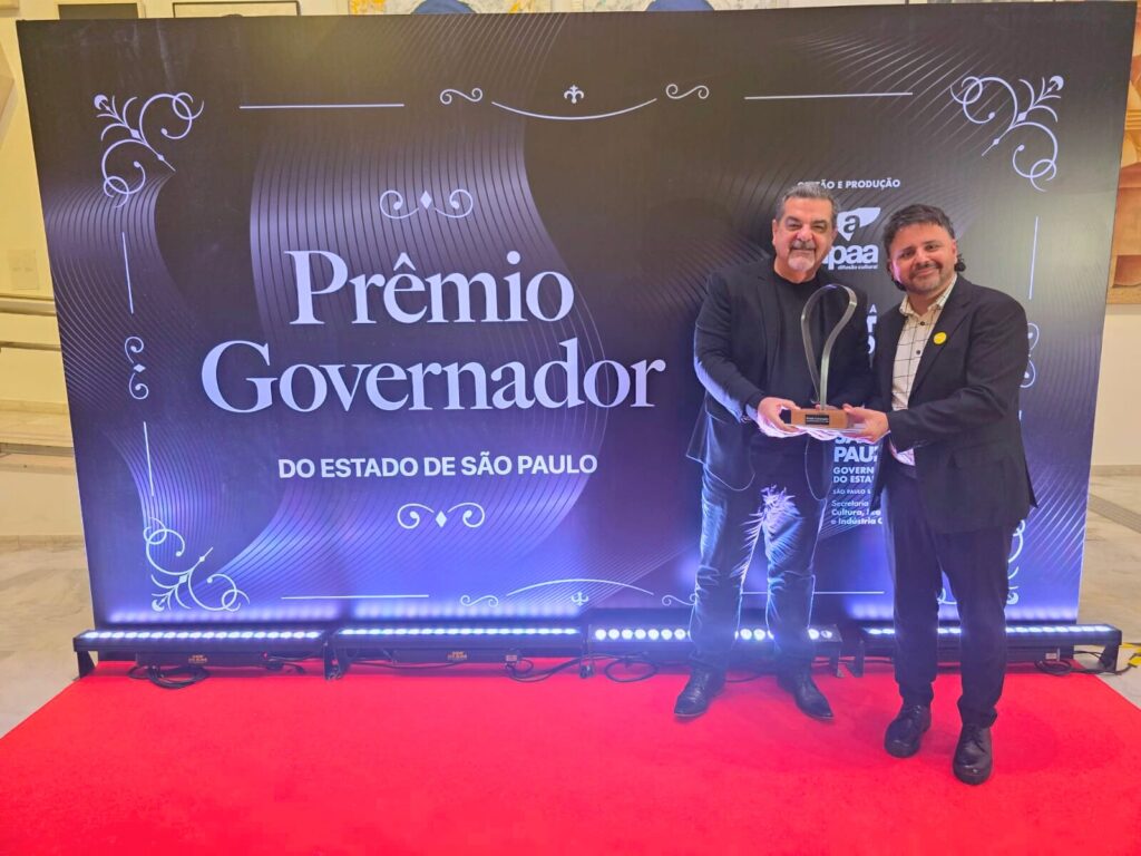 A imagem mostra dois homens em um evento de premiação. Eles estão em pé na frente de um backdrop com a inscrição "Prêmio Governador do Estado de São Paulo". Um dos homens está segurando um troféu. O ambiente parece ser formal, com um tapete vermelho e iluminação profissional. Os homens parecem estar vestidos a rigor, em trajes sociais escuros. A imagem é nítida e bem iluminada, sugerindo uma fotografia profissional de um evento importante. O fundo escuro e elegante contrasta com a iluminação e as cores vibrantes do tapete vermelho e do troféu. A imagem transmite uma sensação de sucesso e formalidade.
