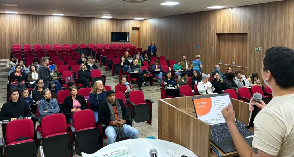 A imagem mostra uma sala de conferências com muitas pessoas sentadas em cadeiras vermelhas. Há um palestrante no palco, usando um microfone e olhando para a plateia. A sala está bem iluminada e organizada. No palco, há um laptop e um cavalete com um programa da conferência. Há um grande número de pessoas presentes, sugerindo um evento importante. A decoração é simples e funcional, típica de uma sala de aula ou auditório. A imagem parece ter sido tirada durante uma apresentação ou palestra.