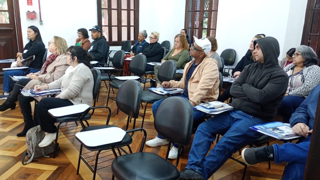 A imagem mostra uma sala de aula com várias pessoas sentadas em carteiras individuais. As carteiras estão dispostas em filas, e as pessoas parecem estar participando de uma palestra ou aula. Há uma mistura de homens e mulheres de diferentes idades, e a maioria deles está vestindo roupas casuais. A sala tem paredes brancas, um piso de madeira e algumas janelas que deixam entrar luz natural. Há também um extintor de incêndio visível na parede. A atmosfera parece calma e atenta. As pessoas parecem estar prestando atenção a algo que está acontecendo na sala.
