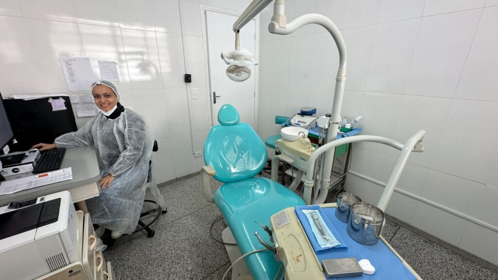 A imagem mostra uma clínica dentária ou consultório odontológico. Uma profissional de saúde, usando avental e máscara, está sentada em frente a um computador, aparentemente trabalhando em alguns papéis ou registros. Ao fundo, há uma cadeira odontológica turquesa, equipamentos odontológicos padrão (como uma unidade de luz e uma bandeja de instrumentos) e uma pequena mesa auxiliar. O ambiente parece limpo e bem organizado, com paredes de azulejos brancos. A iluminação é suave e a imagem é nítida. Em resumo, a cena retrata um ambiente clínico típico de um consultório odontológico.