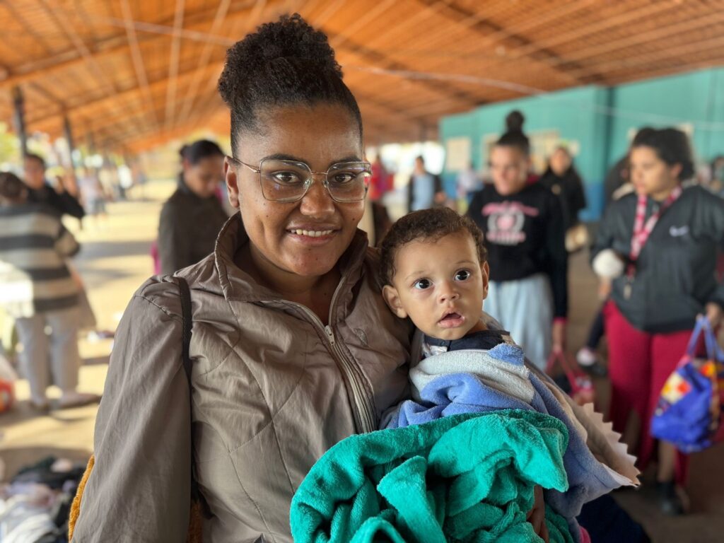 A imagem mostra uma mulher negra com óculos sorrindo e segurando um bebê envolto em um cobertor verde-água. A mulher está usando uma jaqueta marrom-acinzentada e o bebê parece ter cerca de um ano de idade. Ao fundo, há outras pessoas desfocadas, sugerindo um ambiente público como uma estação de ônibus ou abrigo. A atmosfera geral é calma e acolhedora, embora haja um leve tom de melancolia na expressão da mulher. A qualidade da imagem é boa, com cores vibrantes e boa iluminação, apesar de ser aparentemente uma foto tirada com um celular. A imagem transmite uma sensação de ternura e cuidado materno, contrastando com o ambiente público e ligeiramente caótico do fundo.