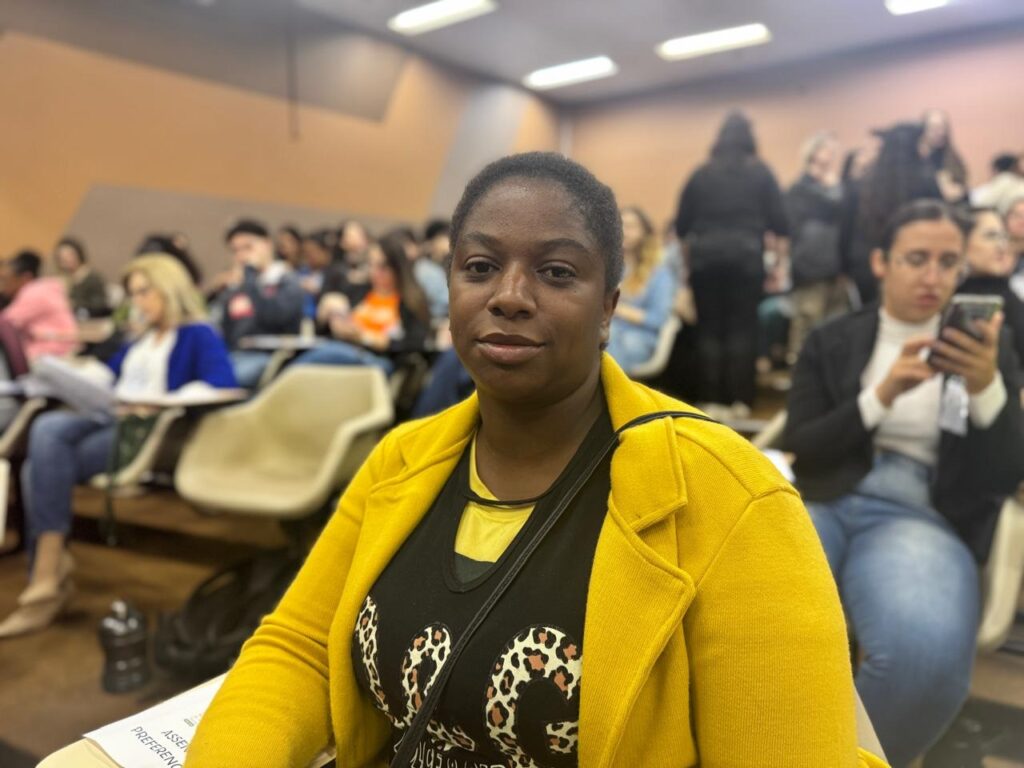 A imagem mostra uma mulher negra de pele escura em primeiro plano, sentada em uma cadeira em uma sala de aula. Ela está usando um casaco amarelo e uma camiseta preta com um padrão de leopardo. Ela parece estar olhando diretamente para a câmera. Há muitas outras pessoas na sala de aula, sentadas em fileiras de cadeiras, mas elas estão fora de foco. A sala de aula parece ser um espaço de aprendizagem moderno, com iluminação artificial e paredes de cor bege. No canto inferior esquerdo da imagem, há uma folha de papel com texto ilegível. A imagem como um todo é bem iluminada e nítida.