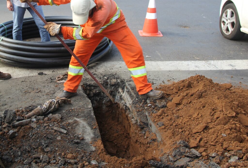 A imagem mostra um trabalhador em um espaço urbano, utilizando roupas de segurança laranjas e uma tampa de proteção na cabeça. Ele está usando uma barra para remover a terra de um buraco no chão. Ao fundo, podem ser vistos cones de sinalização e um rolo de cabo.