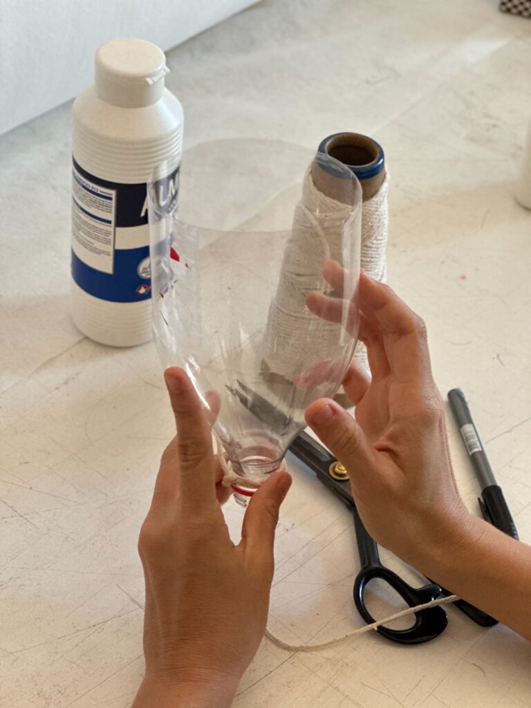 A imagem mostra um close-up de duas mãos segurando uma garrafa de plástico cortada e moldada em forma de funil. Um carretel de barbante está ao lado, e uma tesoura e uma garrafa de cola branca estão visíveis ao fundo. A superfície de trabalho é branca e parece ser uma mesa. A cena sugere um projeto de artesanato ou uma atividade de bricolage. A iluminação é natural e suave. As mãos parecem pertencer a uma pessoa de pele clara. Os detalhes são nítidos e a imagem está bem focada.